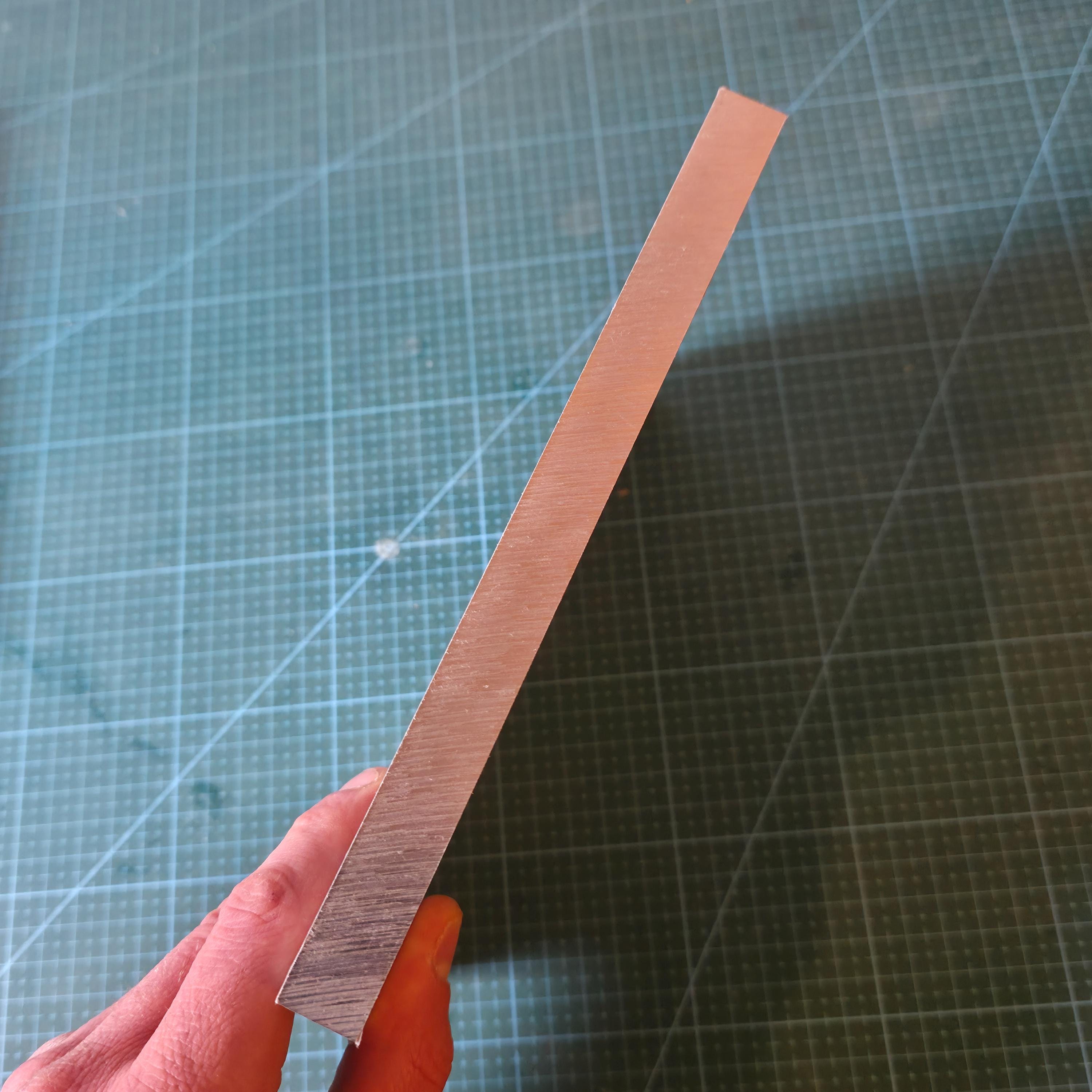 6061 Aluminum Plate edge view held in hand over a blue gridded cutting mat, showing plate thickness and brushed surface finish, raw CNC stock for milling, drilling, tapping, and custom part fabrication.
