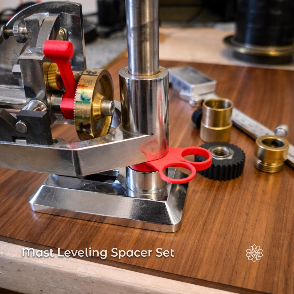 Close-up of a Vevor faceting machine mast mounted on a metal base, shown on a wooden workbench. A red plastic spacer is installed between the mast base and the machine platform, illustrating height adjustment and leveling. Additional brass and plastic components are visible in the background. On-image text reads: “Mast Leveling Spacer Set.” Image represents a precision adjustment accessory used to correct mast alignment on faceting machines.