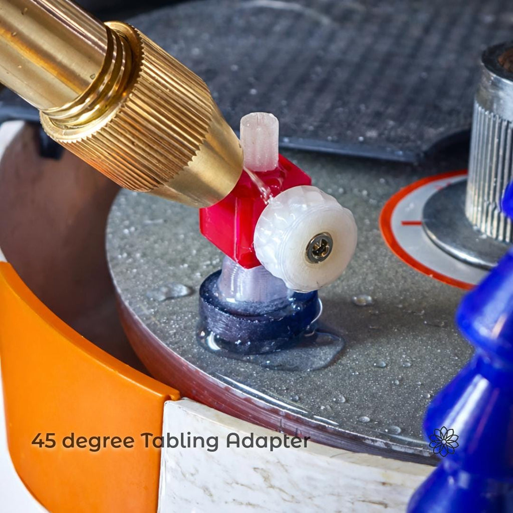 Macro photo of a gemstone dop mounted on a faceting machine lap with a red and white plastic adapter installed beneath the dop. Water is visible on the spinning lap surface, and a brass dop holder is angled toward the stone. An orange basin wall and blue coolant hose appear in the background. On-image text reads: “45 degree Tabling Adapter.” Image shows controlled table cutting setup on a faceting machine.