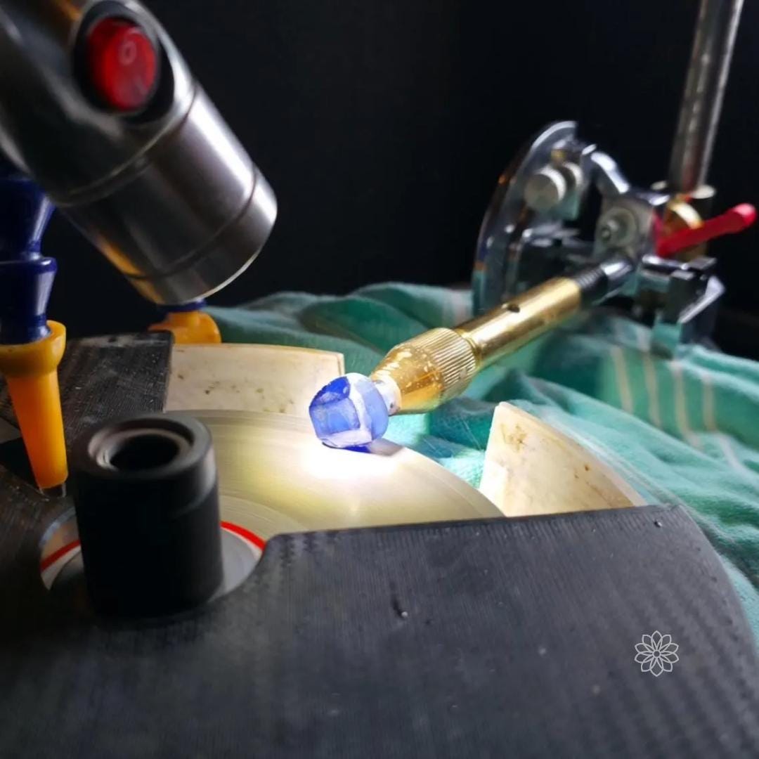 Faceting machine cutting setup with a blue rough sapphire gemstone mounted on a brass dop stick touching a spinning lap under a work light. The faceting head and quill assembly are visible, demonstrating stone shaping during early faceting preparation.