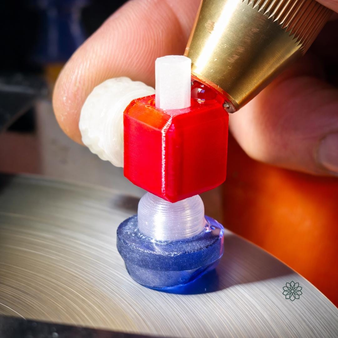 Macro view of the Aetherium 45-degree tabling adapter holding a blue sapphire gemstone dop against a spinning faceting lap. The red polymer body stabilizes the dop while a brass faceting handpiece presses the stone into the diamond lap for cutting a flat surface. Coolant water and lap texture are visible, illustrating controlled table cutting on a lapidary faceting machine.