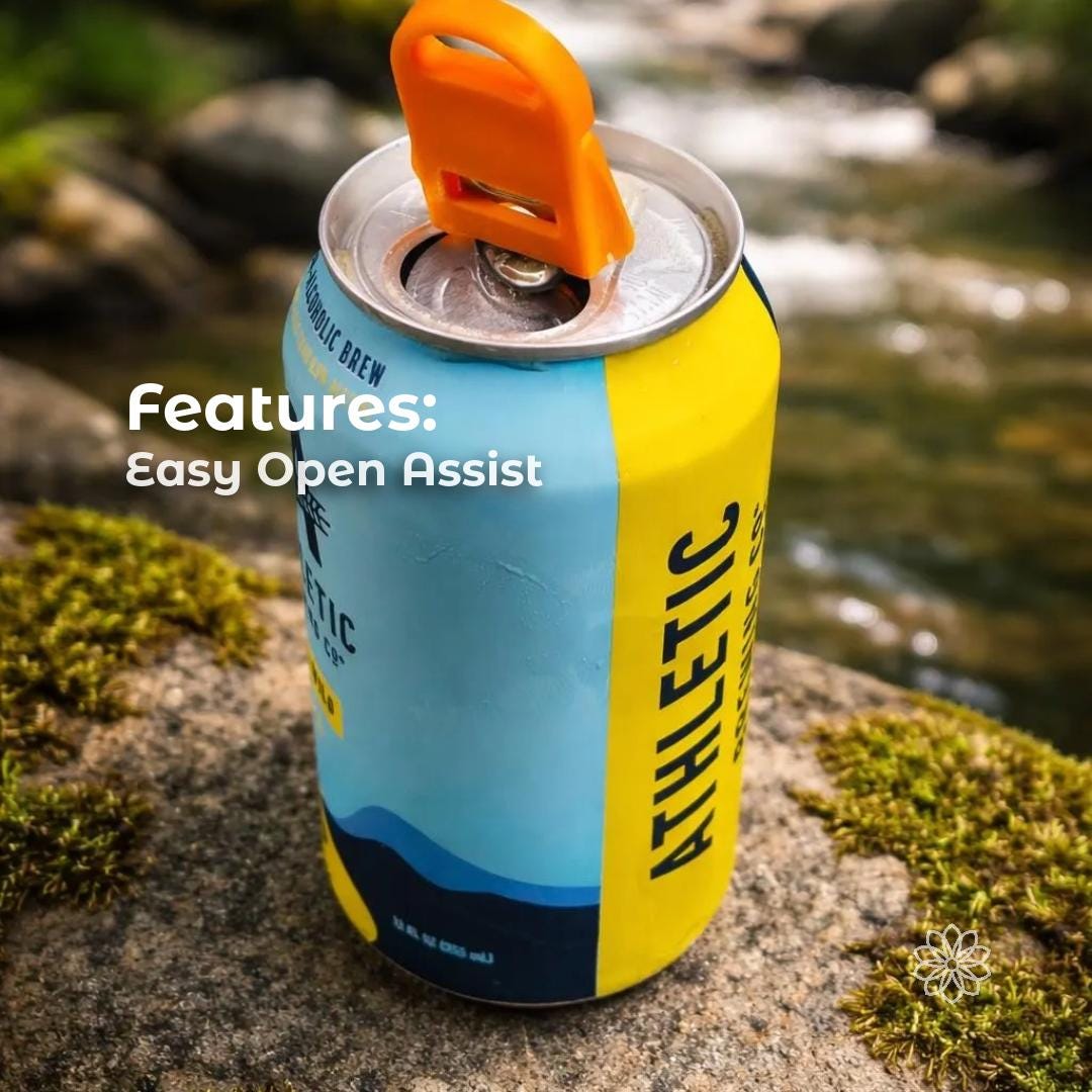 Close-up of an orange Aetherium Can Guard tool lifting the pull tab on a beverage can beside a rocky creek outdoors. The compact camping accessory acts as an easy-open assist for soda or beer cans while protecting fingers from sharp tabs. Overlay text reads “Features: Easy Open Assist,” highlighting the tool’s use for outdoor drinks at campsites, beaches, or picnics.