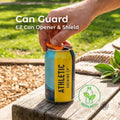 Hand opening a beverage can using the Aetherium Can Guard EZ can opener and drink shield attached to an aluminum drink can on a wooden picnic table outdoors. The orange camping can tool lifts the pull tab without finger strain and rotates to cover the opening, helping keep bugs and debris out during camping, picnics, or outdoor gatherings. Overlay text reads “Can Guard – EZ Can Opener & Shield” with a bioplastic material icon.