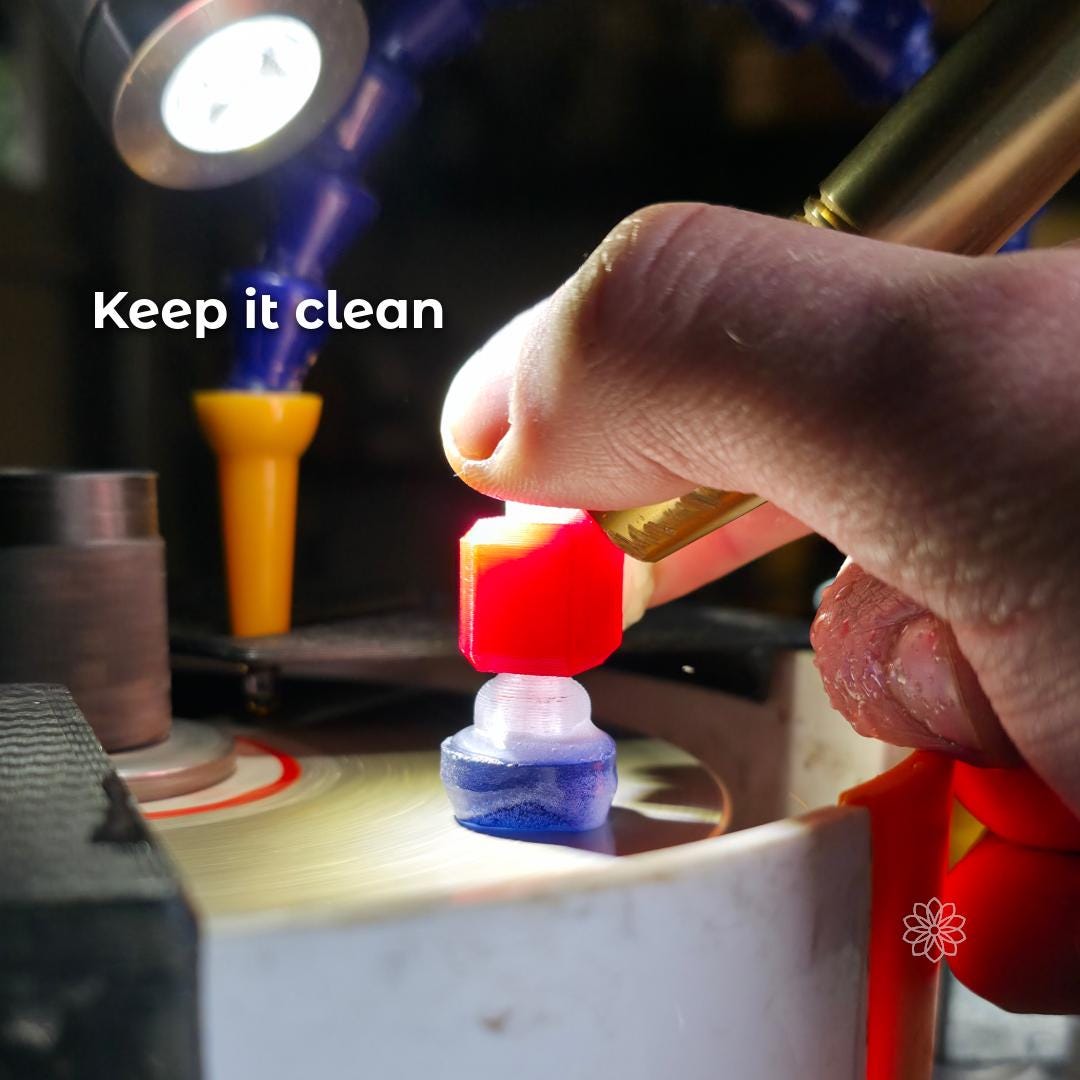 Hand holding a brass faceting handpiece over a spinning lap disk while cutting a red gemstone on a dop inside a faceting machine basin. The scene shows the messy cutting environment the lap disk cover is meant to keep cleaner by containing slurry and spray. Overlay text reads “Keep it clean.”
