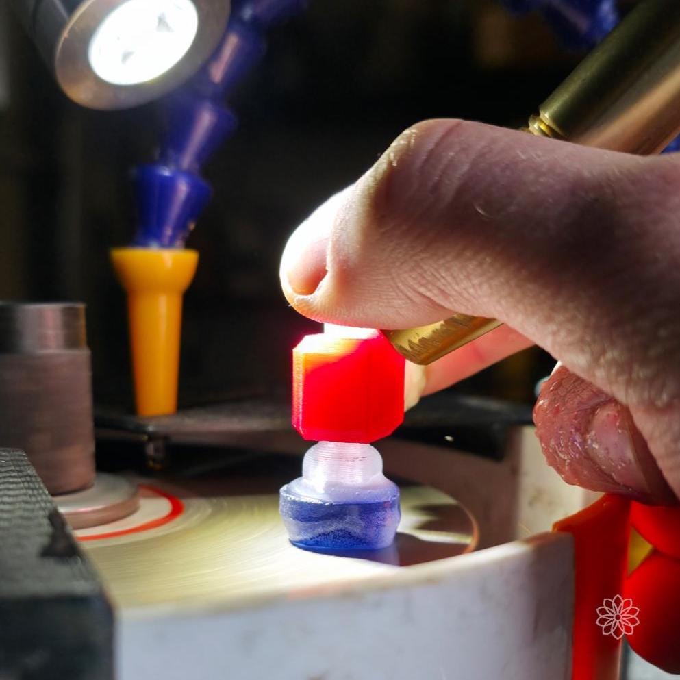 Hand positioning a sapphire gemstone dop into the Aetherium 45-degree tabling adapter on a faceting lap machine. The red polymer adapter provides a fixed angle reference while the user guides the stone across the rotating lap under a work light. Image demonstrates controlled table cutting and stable geometry using a 6 mm dop faceting system.