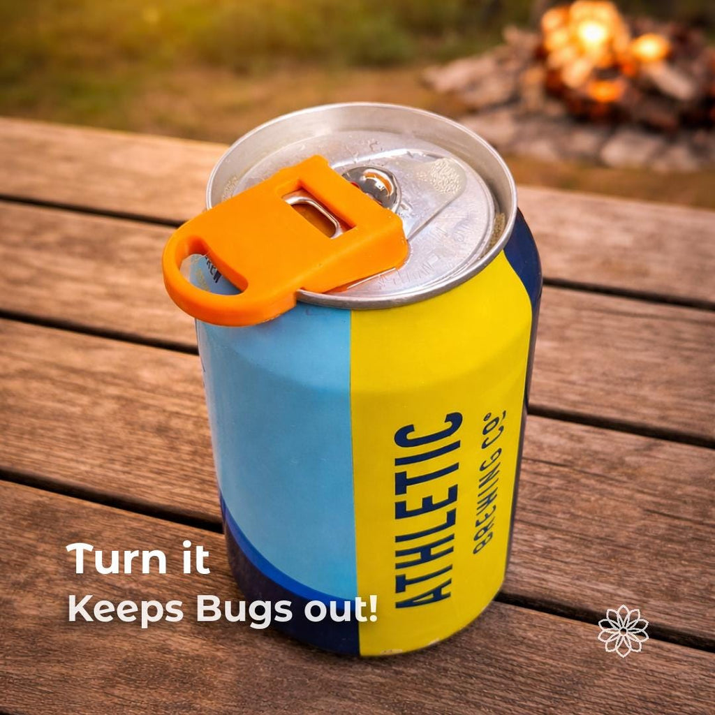 Beverage can with the orange Aetherium Can Guard rotated over the opening to act as a drink shield while sitting on a wooden picnic table near a campfire. The camping accessory covers the can opening to help keep bugs and debris out while drinking outdoors. Overlay text reads “Turn it Keeps Bugs Out!” demonstrating the rotating cover function.