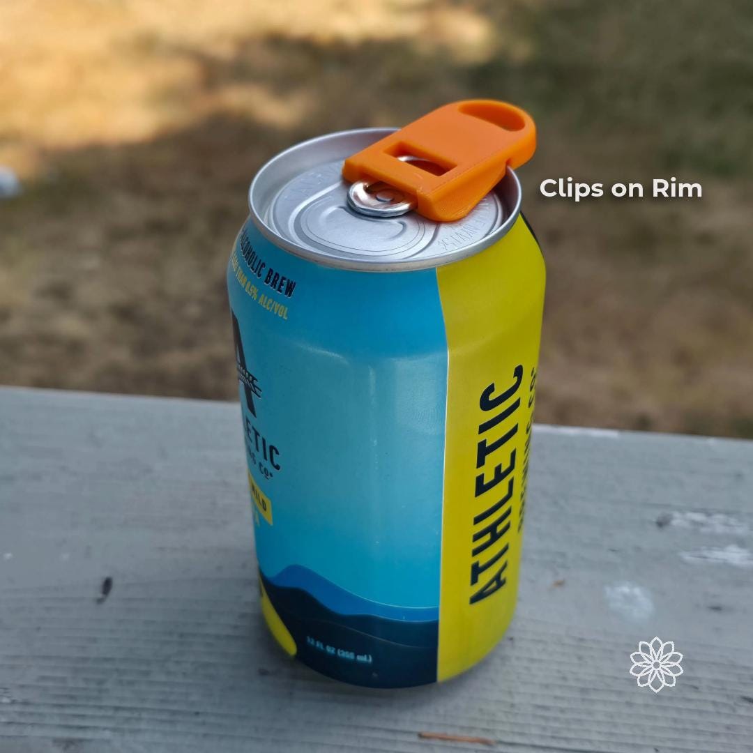 Aetherium Can Guard EZ can opener clipped onto the rim of a standard aluminum beverage can. The orange camping drink accessory sits securely over the pull tab and rim, allowing the user to open the can and rotate the tool to shield the drinking opening from bugs or dirt. Overlay text reads “Clips on Rim,” demonstrating the simple attachment design.