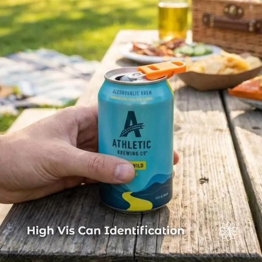 Hand holding a beverage can with the Aetherium Can Guard opener and drink shield attached on a picnic table beside food and drinks outdoors. The bright orange camping tool sits on the pull tab, allowing the user to open the can easily and rotate the cover to protect the drink opening from insects or debris during outdoor meals. Overlay text reads “High Vis Can Identification.”