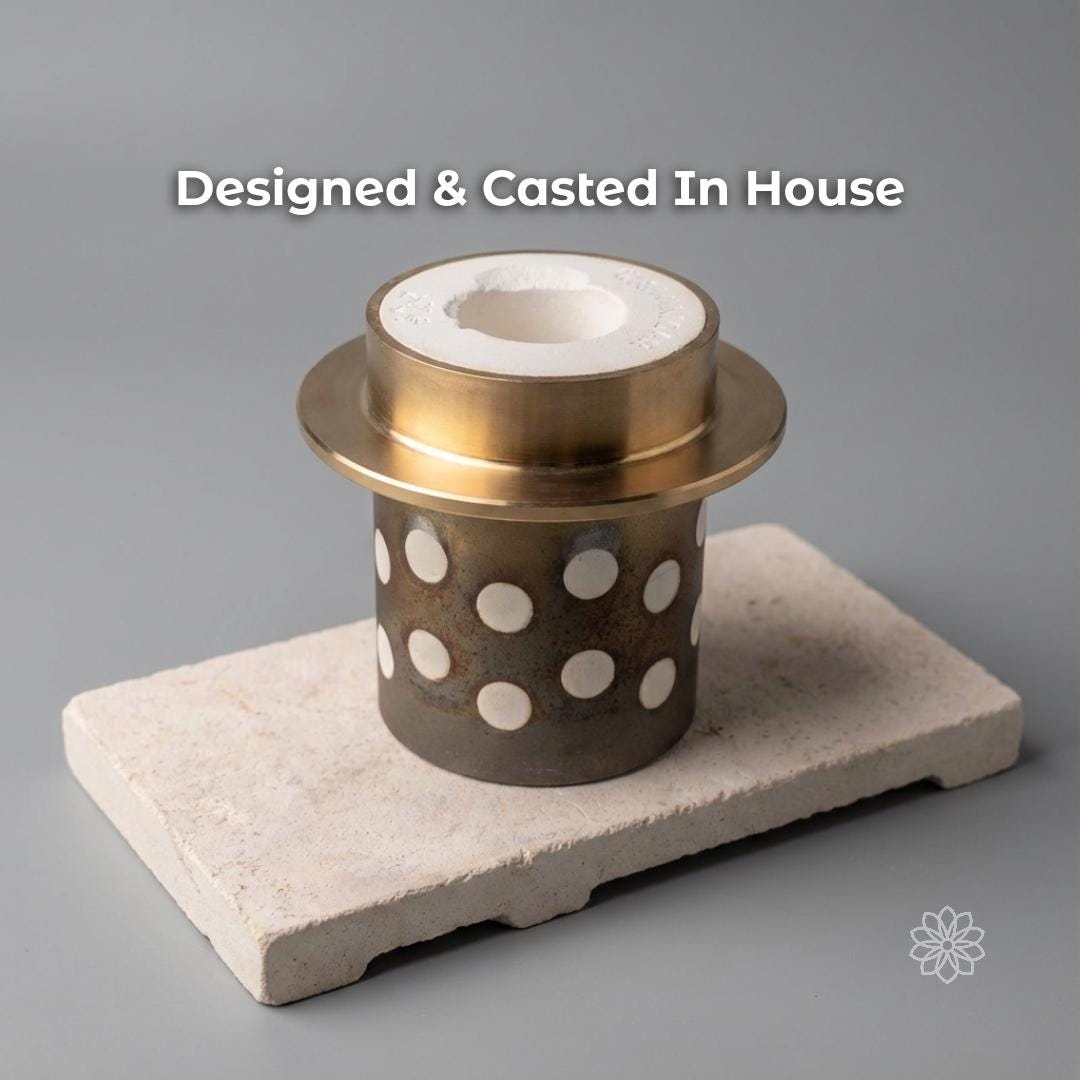 Perforated metal casting flask with a white investment core sits on a ceramic block against a plain background, representing the in-house casting process used to make Aetherium jewelry. Visible text says “Designed & Casted In House.”