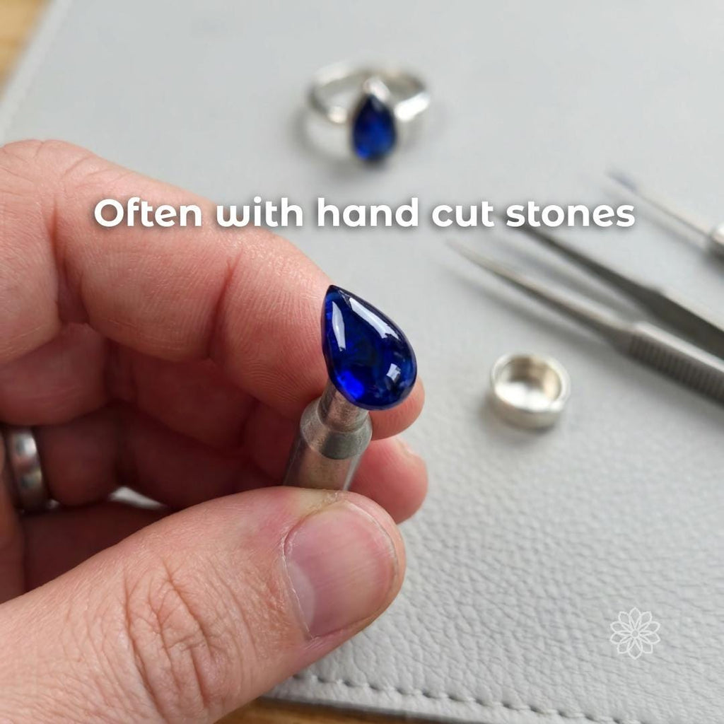 Hand holding a deep blue pear-shaped sapphire gemstone mounted on a dop stick during jewelry work, with ring components and tweezers visible on a work surface. This image shows the hand-cut stone process used in custom jewelry creation. Visible text says “Often with hand cut stones.”