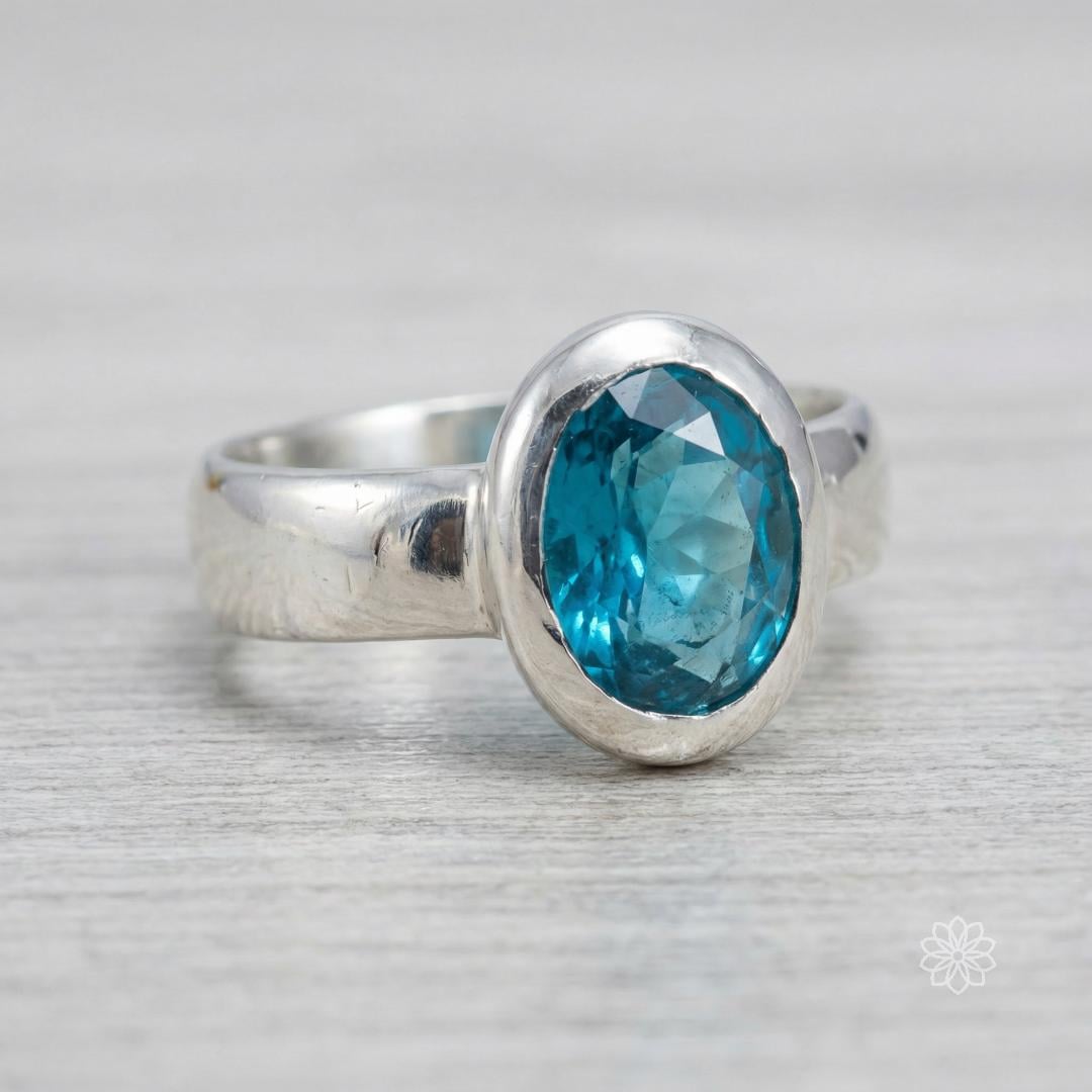 Close-up of a sterling silver bezel-set oval sapphire ring showing the teal blue faceted stone, surface reflections, and handmade finish on the band and hammered bezel.