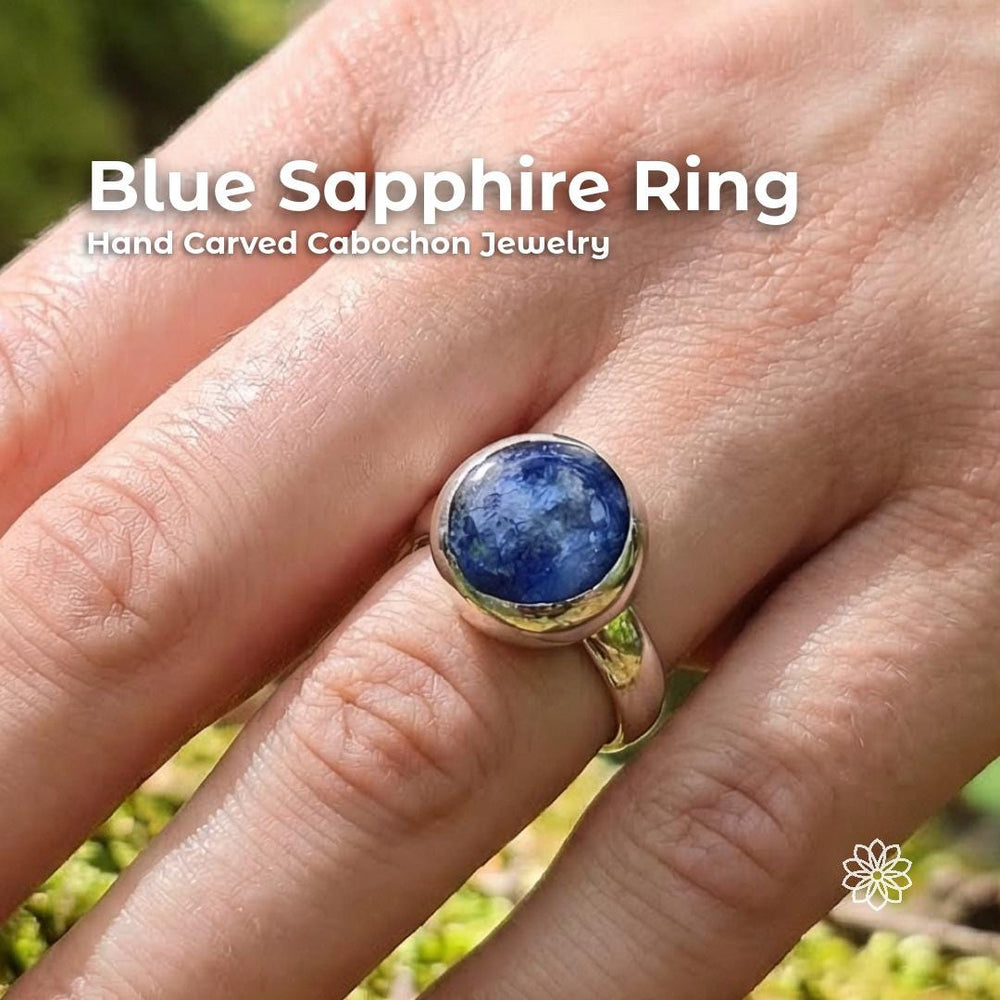 Blue Madagascar sapphire cabochon ring worn on hand outdoors, featuring a deep blue round stone with visible natural inclusions set in a sterling silver bezel. Text reads “Blue Sapphire Ring” and “Hand Carved Cabochon Jewelry.”