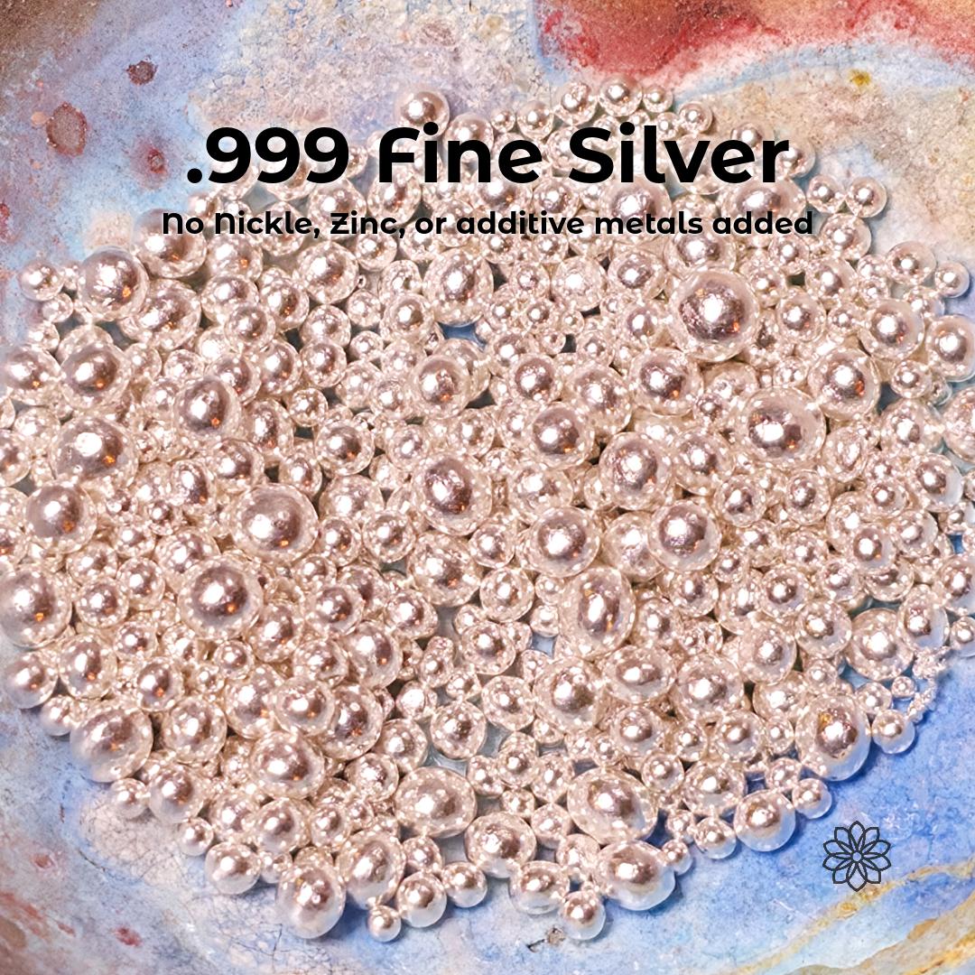 Close-up of .999 fine silver casting grain with smooth reflective spherical beads in multiple sizes on textured surface, text overlay reads “.999 Fine Silver No nickel, zinc, or additive metals added,” showing high purity silver material for jewelry casting and fabrication