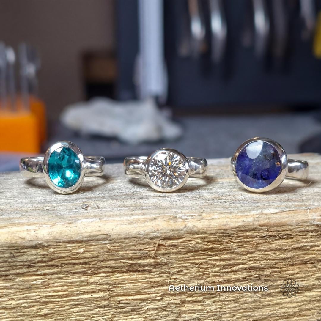 Three sterling silver bezel-set rings displayed side by side on wood, featuring different stones including green, clear, and deep blue gemstones. This image shows variation in stone color and style within the same modern ring design. Visible text says “Aetherium Innovations.”