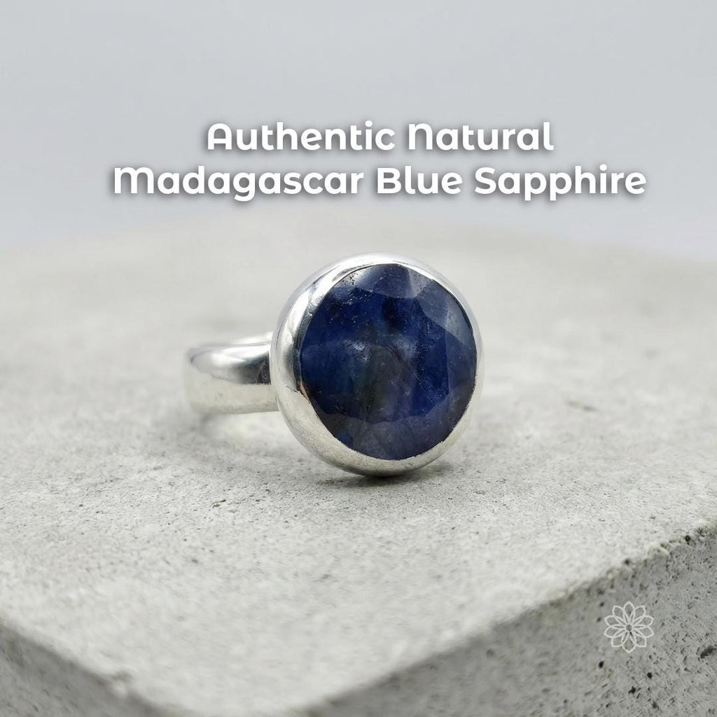 Sterling silver bezel-set blue sapphire cabochon ring on a stone surface, showing a deep blue natural stone with cloudy inclusions and polished dome. Text reads “Authentic natural madagascar Blue Sapphire.”