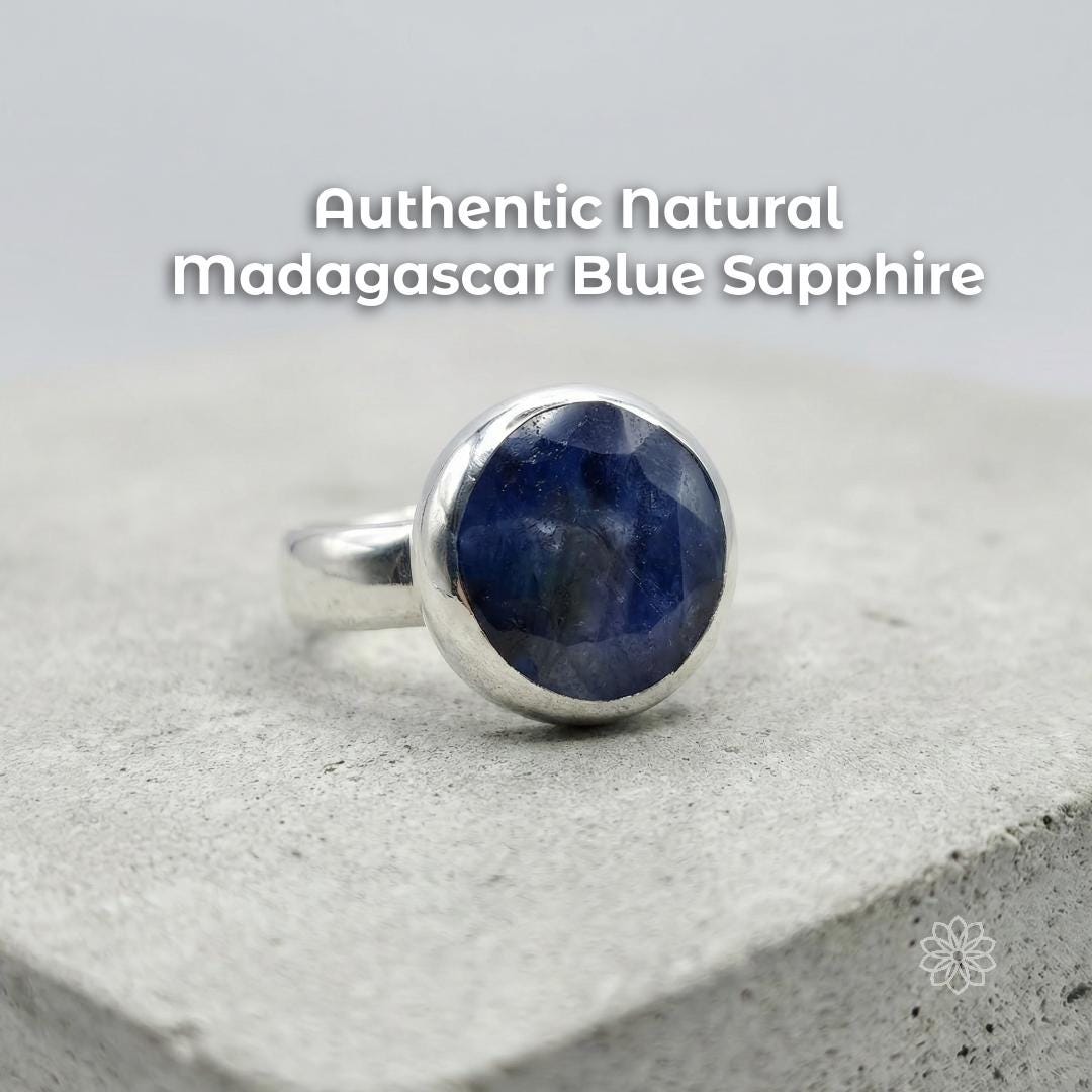 Sterling silver bezel-set blue sapphire cabochon ring on a stone surface, showing a deep blue natural stone with cloudy inclusions and polished dome. Text reads “Authentic natural madagascar Blue Sapphire.”