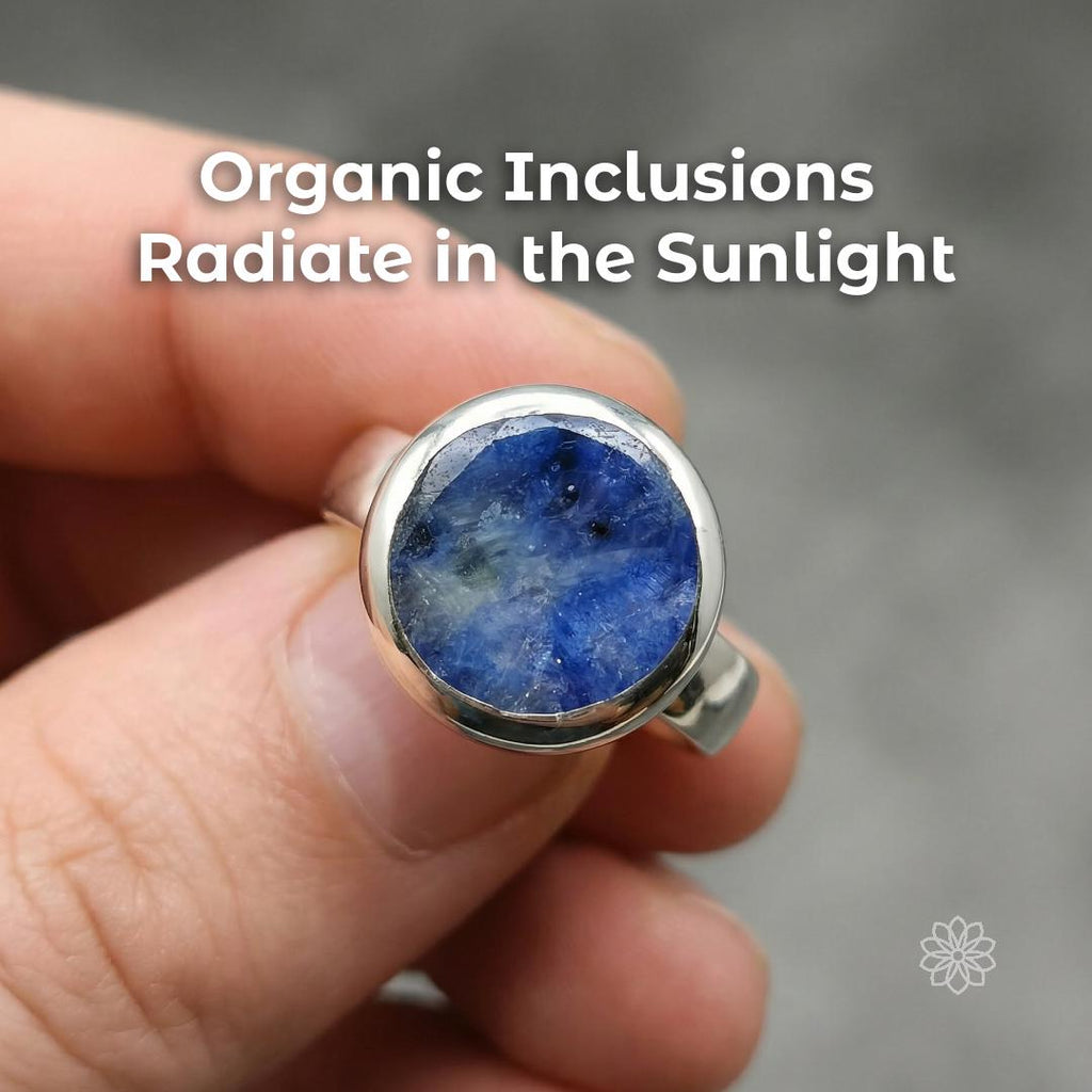 Close-up of a blue sapphire cabochon showing natural inclusions and cloudy internal patterns under light, set in a smooth sterling silver bezel. Text reads “Organic Inclusions Radiate in the Sunlight.”