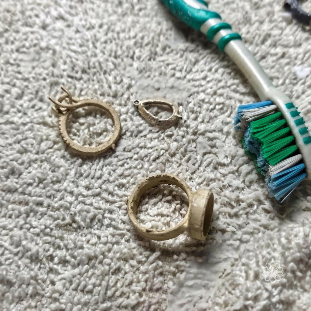 Raw ring components and casting remnants on a work surface next to a brush, showing unfinished metal parts used in the jewelry making process for handmade rings.