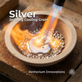 silver jewelry casting grain melting in a ceramic crucible under a torch flame, bright orange heat surrounding liquid silver pool with solid beads around the edge, text overlay reads “Silver Jewelry Casting Grain” and “Aetherium Innovations,” showing real casting process and metal behavior