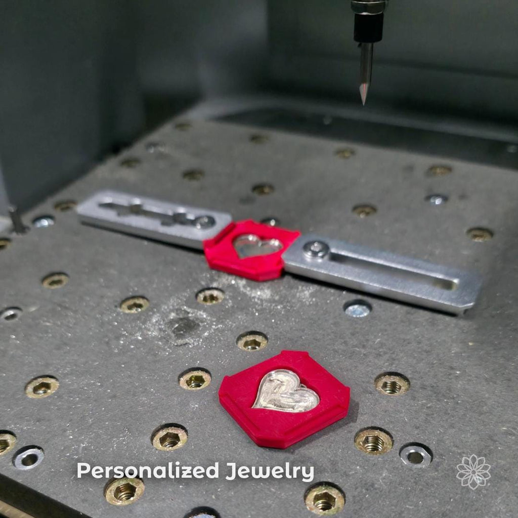 CNC workspace with small heart-shaped metal insert set into a red fixture on a precision bed. Overlay text reads “Personalized Jewelry.” Demonstrates ability to machine custom jewelry components, engraved inserts, and small decorative pieces with fine detail and controlled positioning.