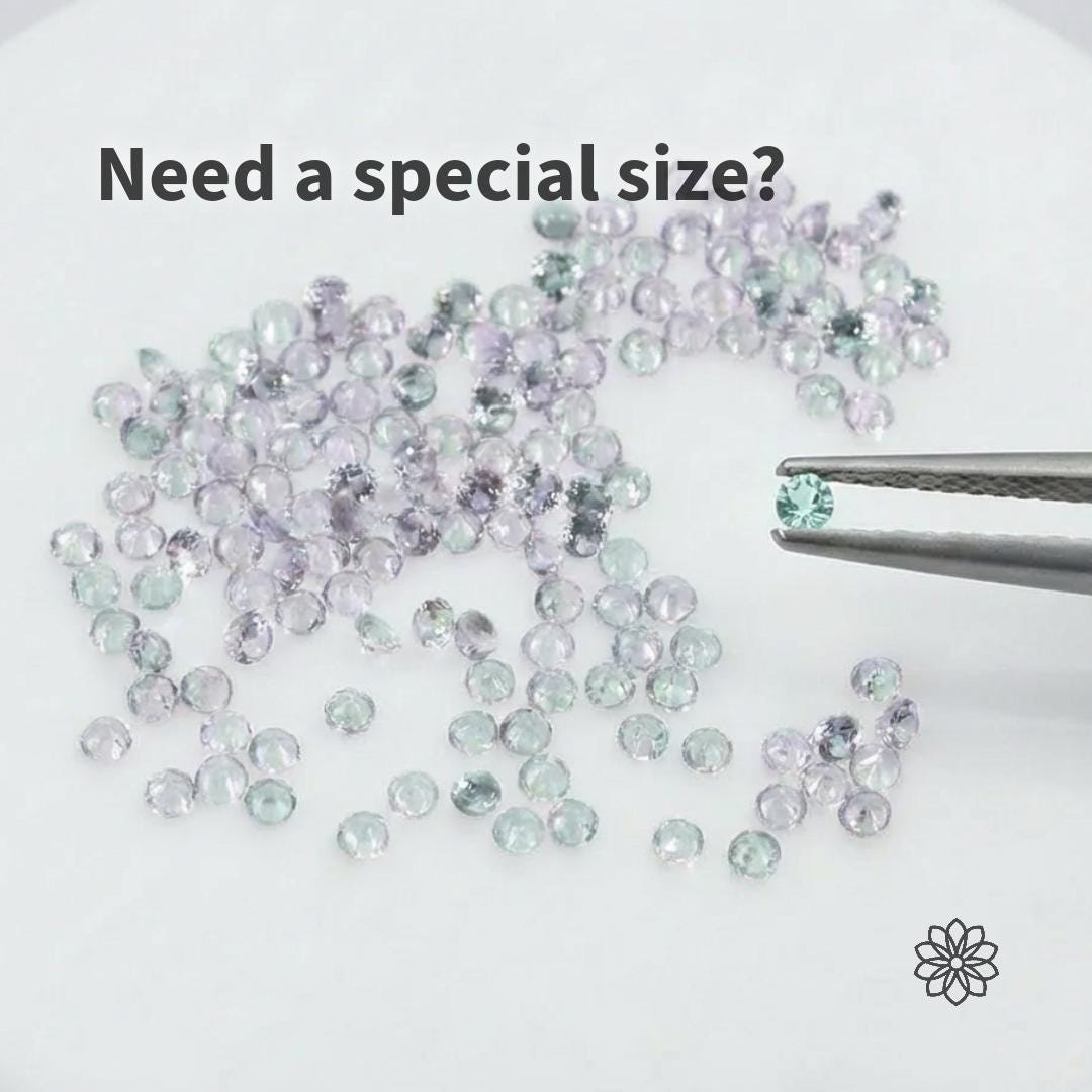 Loose calibrated alexandrite gemstones in pale mint green and soft lavender hues scattered on a white surface, with metal tweezers holding a single round stone for scale. Image demonstrates alexandrite color shift and supports custom sizing options for jewelry makers and precision stone selection.