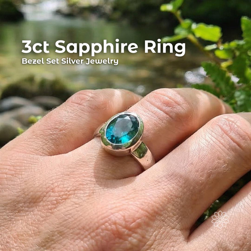 Hand wearing a 3ct teal blue sapphire ring set in sterling silver bezel, photographed outdoors near a flowing creek with natural greenery. Oval cut stone shows deep blue and teal color variation under sunlight. Handmade silver band with organic finish, worn on finger to show real scale and everyday wear.