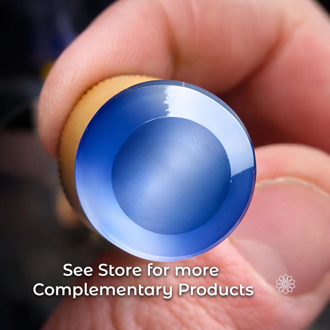Close-up of a finished circular Sapphire gemstone window viewed straight on, showing a smooth, polished surface with a blue gradient reflection. The stone is mounted on a dop stick held between fingers. On-image text reads: “see store for more complementary products.” Image illustrates the surface quality achievable through controlled faceting workflows.