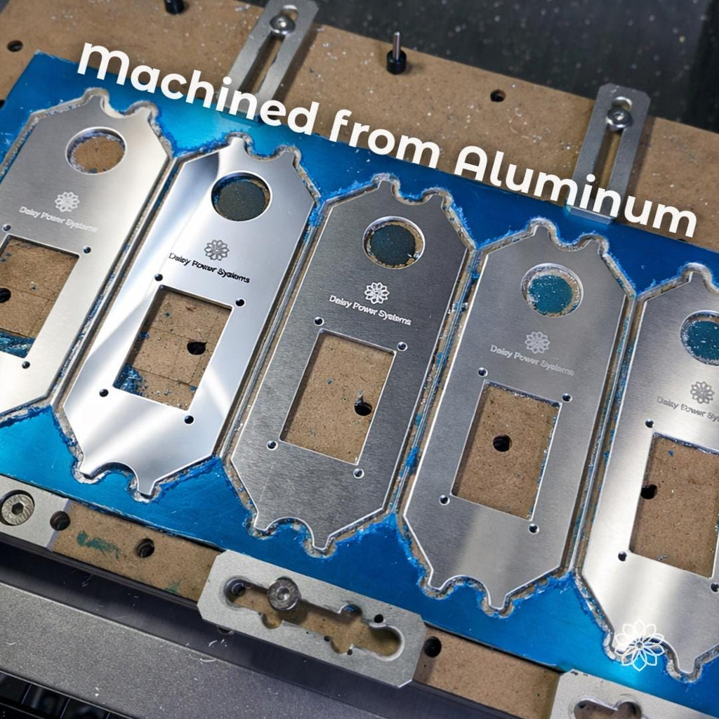 Text reads “machined from Aluminum.” Multiple Daisy Power Systems switch panel faceplates are shown on a CNC fixture with blue protective film, engraved logos, mounting holes, and unfinished machined edges. This image shows the aluminum construction and in-house machining process before finishing and assembly.