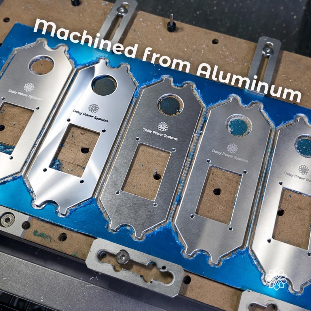 Text reads “machined from Aluminum.” Multiple Daisy Power Systems switch panel faceplates are shown on a CNC fixture with blue protective film, engraved logos, mounting holes, and unfinished machined edges. This image shows the aluminum construction and in-house machining process before finishing and assembly.