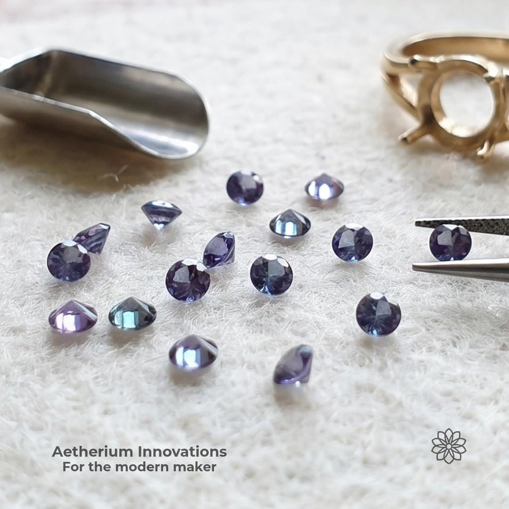 Loose 2mm Czochralski alexandrite stones arranged beside tweezers, a metal scoop, and an unfinished ring setting on a white jeweler’s surface. The blue-violet and teal stones are framed as calibrated lab-grown chrysoberyl melee for modern makers building rings, accents, and small-batch jewelry.
