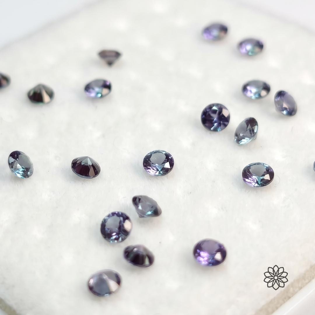 Close view of loose 2mm pulled alexandrite stones on a soft white pad, shown face-up and pavilion-up to reveal cut depth, polish, and color shift. The stones move through purple-violet, teal, and muted purple tones, helping jewelry makers evaluate optical character beyond generic lab alexandrite listings.