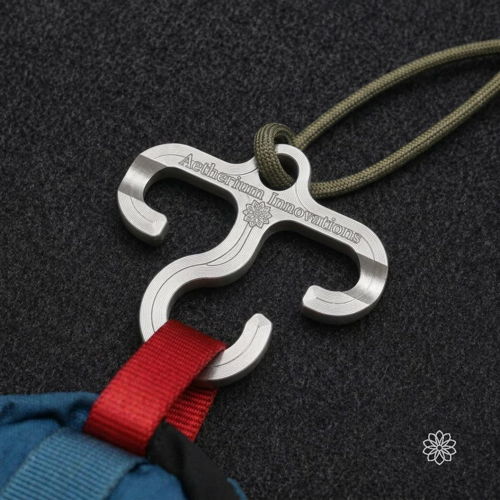 Machined aluminum Paracord Hook Mini attached to green cord and holding red backpack webbing against a dark textured surface. Shows how the compact three-hook design can help hang, stage, or organize lightweight gear from paracord, pack loops, camp lines, and outdoor setups.