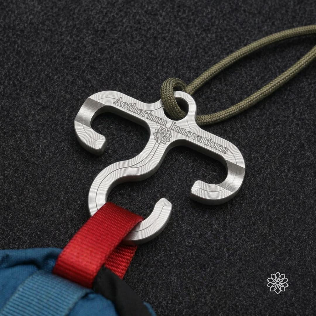 Machined aluminum Paracord Hook Mini attached to green cord and holding red backpack webbing against a dark textured surface. Shows how the compact three-hook design can help hang, stage, or organize lightweight gear from paracord, pack loops, camp lines, and outdoor setups.