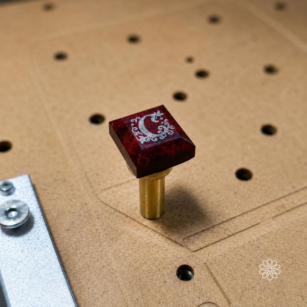 Square red gemstone engraved with an intricate decorative design while mounted on a brass dop stick for CNC machining