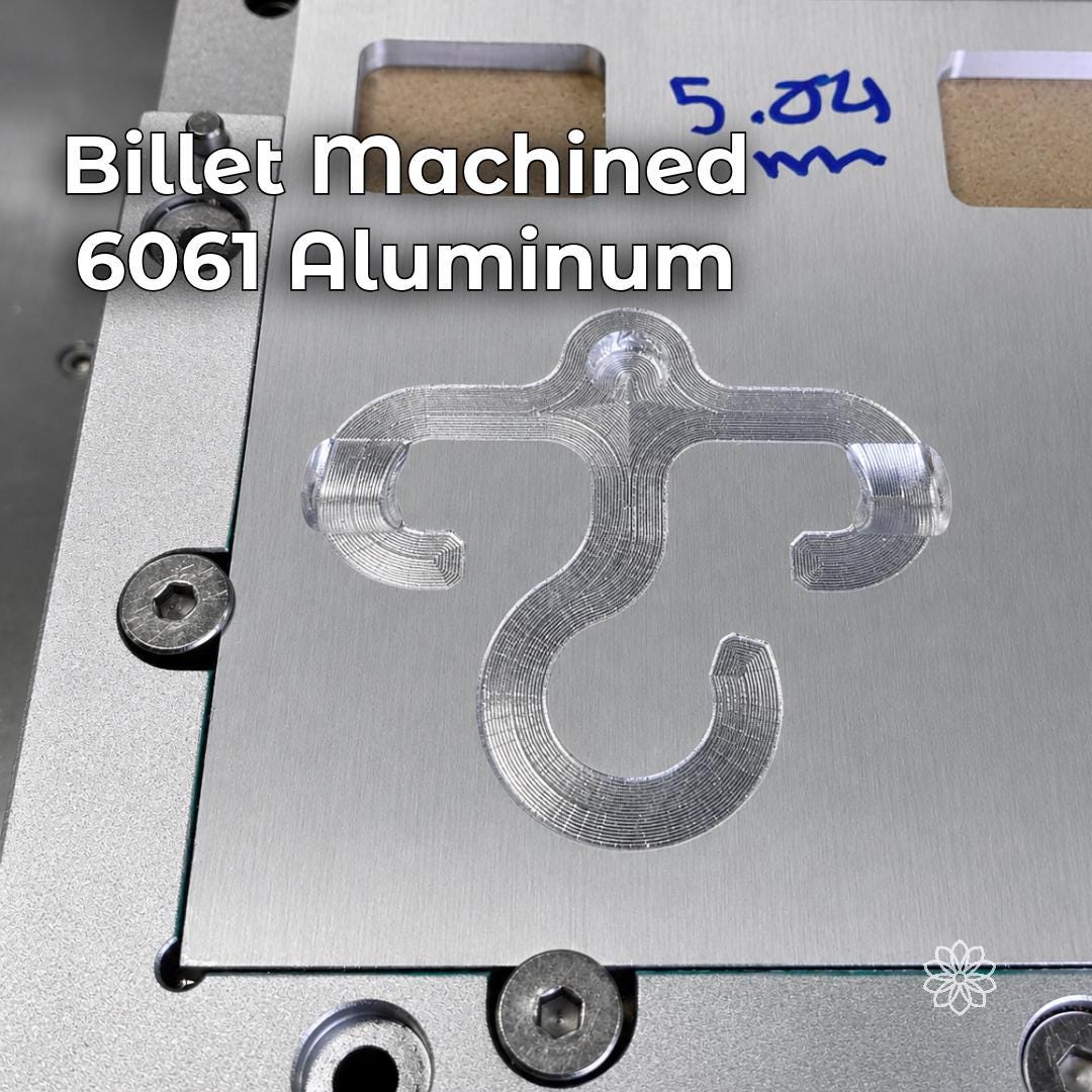 Billet machined 6061 aluminum Paracord Hook Mini shown on a CNC machining plate with visible tool paths and unfinished cut detail. Image highlights the small-batch machined construction, thin aluminum profile, and durable outdoor utility design for EDC, backpacking, and camp gear.