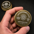 Hand holding a small engraved gold tin of Rockhounder’s Salve, showing the Aetherium logo, Founder’s Harvest batch marking, and pocket-size scale for outdoor, camping, and workshop use.