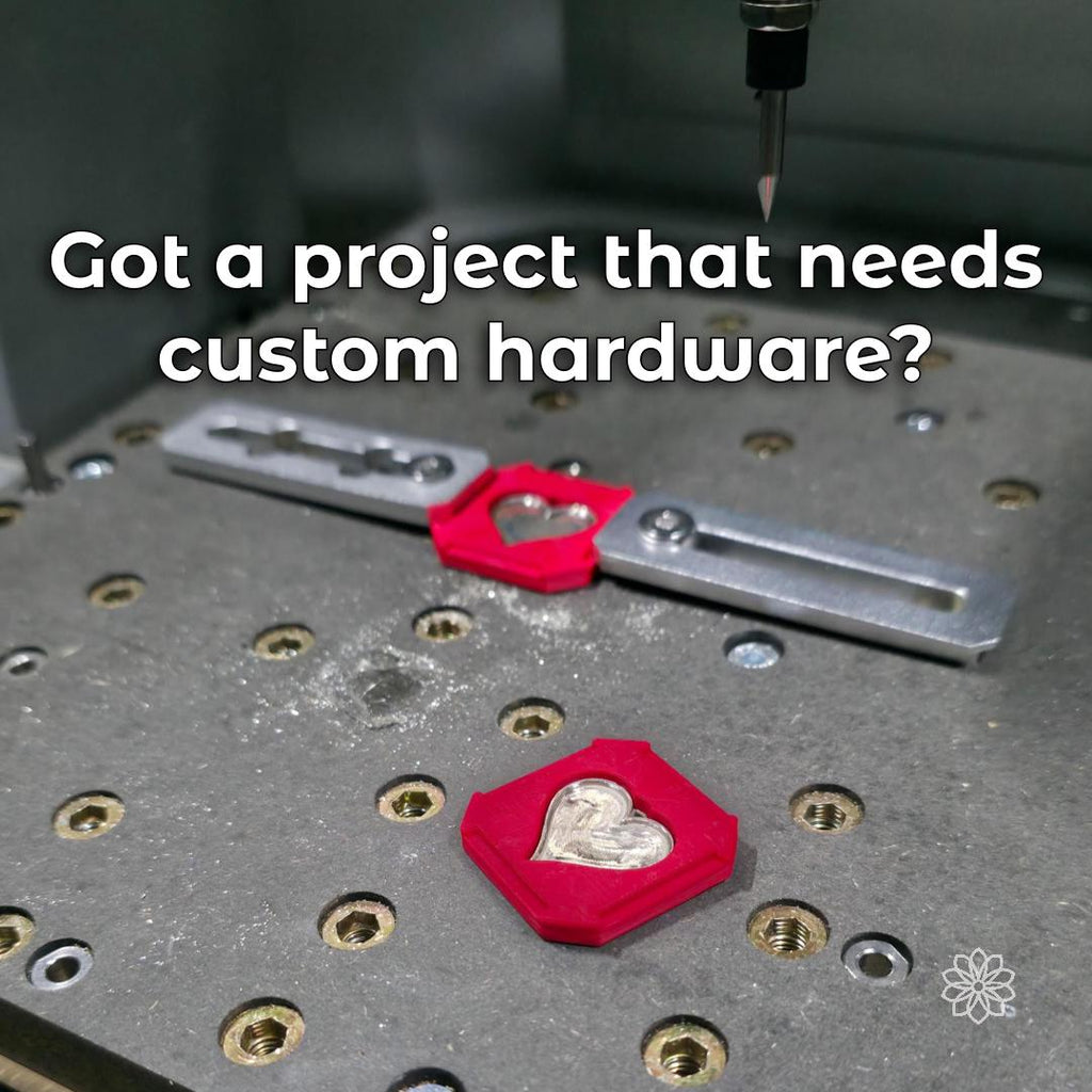 Red 3D-printed CNC fixtures holding two heart-shaped metal pieces on a threaded machine bed beneath an engraving bit, with visible text describing custom hardware projects.