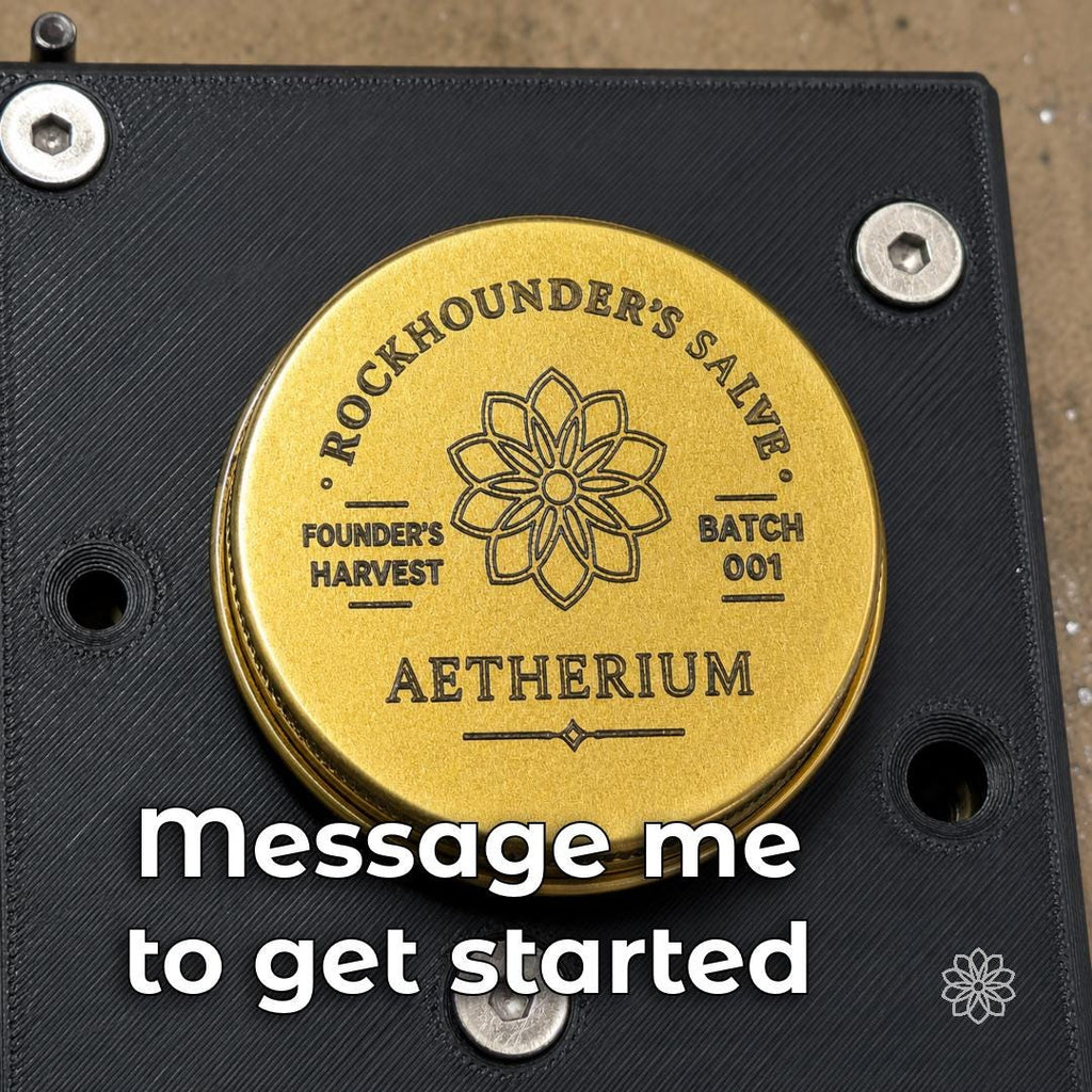 Gold-colored engraved round metal piece secured in a black 3D-printed CNC fixture, with Aetherium markings and custom line engraving visible across the finished circular face.