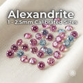 Round-cut lab-grown alexandrite gemstones in calibrated 1 mm to 2.5 mm sizes arranged on white fabric. Faceted stones show vivid pink, lavender, pale blue and green color shifts under changing light. Loose alexandrite stones for jewelry making, pavé settings, halo accents, side stones and precision inlay.