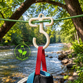 Full-size white bioplastic paracord hook hanging from a green ridgeline beside a wooded river, supporting a red gear strap and camping bag. Lightweight outdoor gear hanger designed for campsite organization, backpacking, bushcraft, overlanding, and hanging lanterns, cookware, clothing, towels, and other camp equipment.