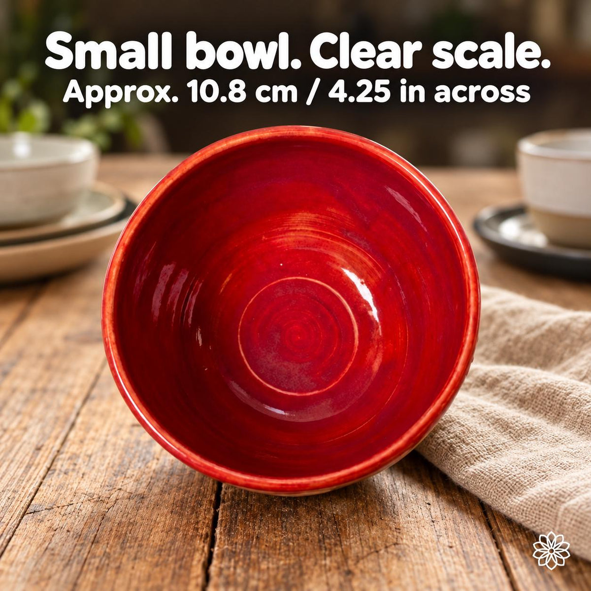 Tilted view inside the red glazed ceramic bowl on wood and linen, with neutral dishware behind. Text: Small bowl. Clear scale. Approx. 10.8 cm / 4.25 in across.